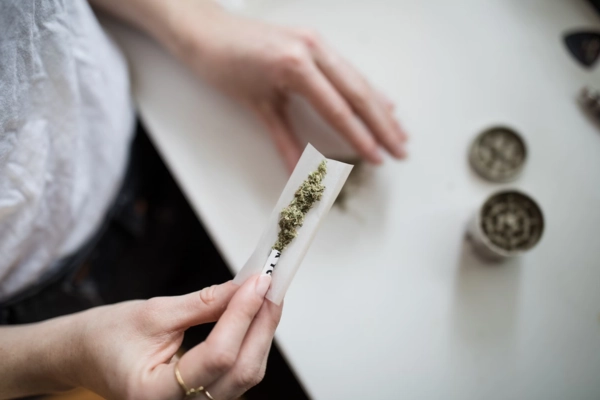 Person rolling cannabis into a paper on a table, with a grinder and loose cannabis in the background