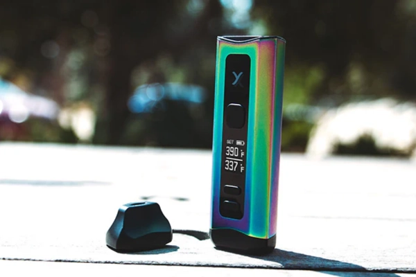 Colorful dry herb vaporizer with temperature display sitting on a wooden table outdoors, sunlight in the background